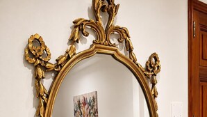 A room with a large ornate gold-framed mirror, a gray upholstered chair, a beige cabinet, and a colorful abstract painting on the wall. - Valentina Rooms – Elegant Stay in Bronte, Pistacchio e Etna. (Bronte)