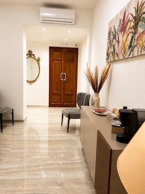 Un área de recepción moderna con suelo de mármol, puerta de madera, espejo, mostrador con cafetera y una planta decorativa.