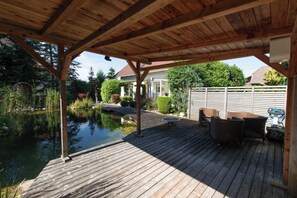 Eine Holzterrasse mit Teich, Gartenmöbeln und überdachtem Bereich.