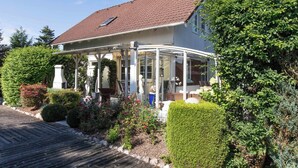 Una casa a due piani con un portico coperto, un tetto in tegole e un giardino curato con diverse piante e arbusti.