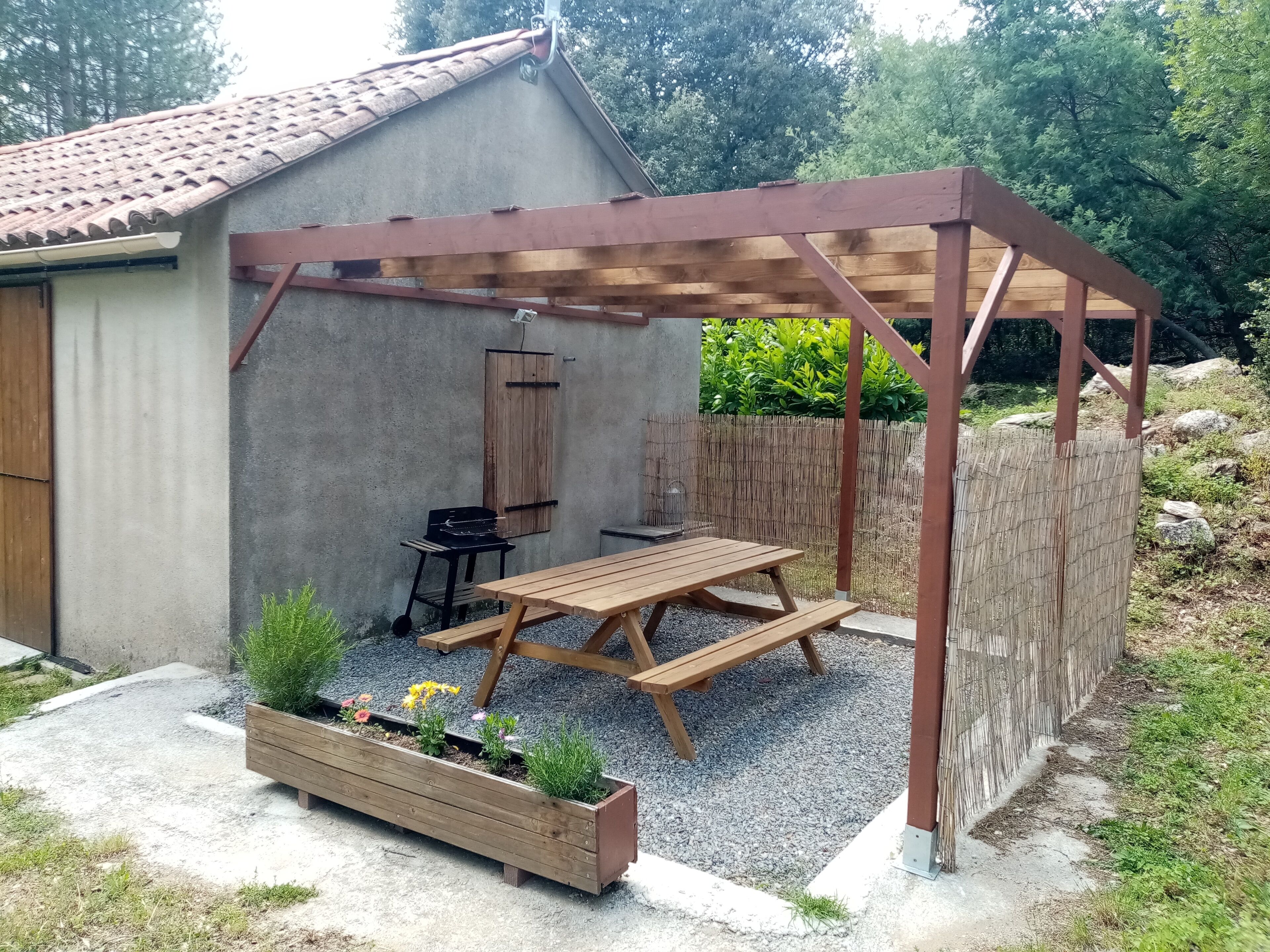 Eine überdachte Terrasse mit Holztisch und -stühlen, einem Grill und einem Blumenbeet mit Pflanzen.