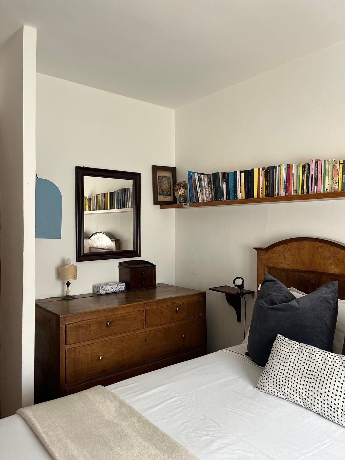 Een slaapkamer met een houten dressoir, nachtkastje, spiegel, boekenkast en een bed met kussens.