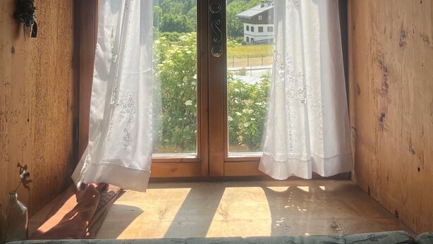 Une pièce avec un plafond en bois, une grande porte-fenêtre coulissante avec des rideaux blancs, et une vue sur une maison et la verdure environnante.