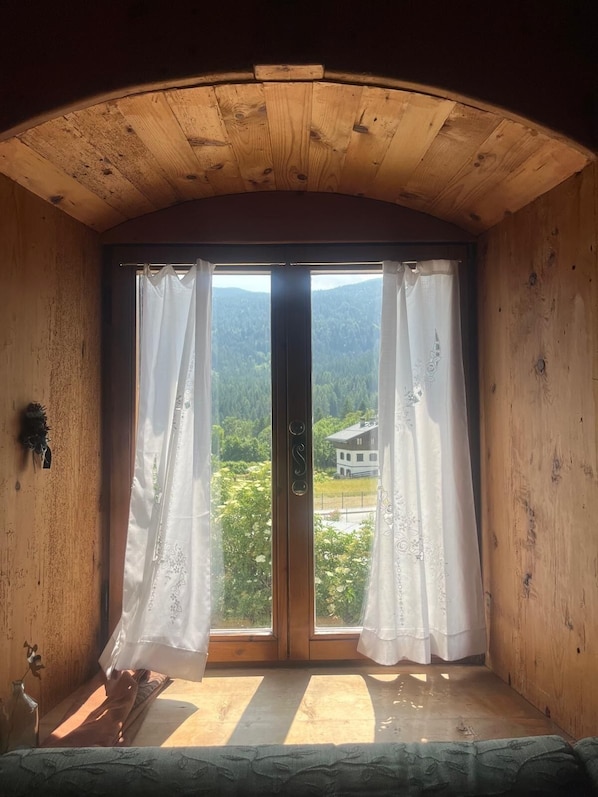 A room with a wooden ceiling, a large sliding glass door with white curtains, and a view of a house and greenery outside.