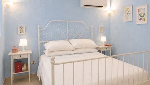 Une chambre avec un lit en métal blanc, deux tables de chevet avec des lampes, un climatiseur fixé au mur et des œuvres d’art florales accrochées aux murs.