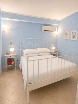 A bedroom with a white metal bed, two bedside tables with lamps, a wall-mounted air conditioner, and floral artwork on the walls. - SMART EMERALD COAST (provincia della gallura nord-est sardegna)