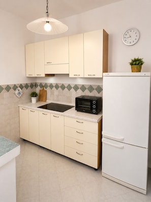 A kitchen with beige cabinets, a black microwave, a white refrigerator, a clock, and a hanging light fixture. - SMART EMERALD COAST (provincia della gallura nord-est sardegna)