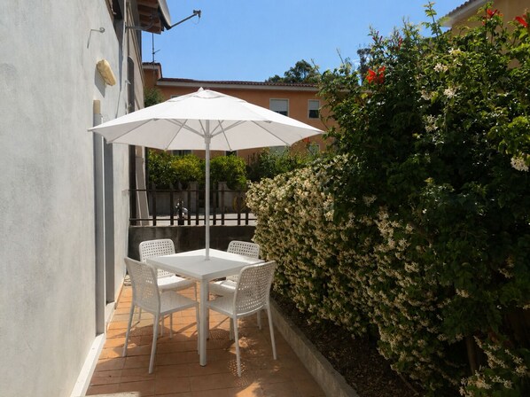A patio with a white table and chairs, an umbrella, and a hedge. - SMART EMERALD COAST (provincia della gallura nord-est sardegna)