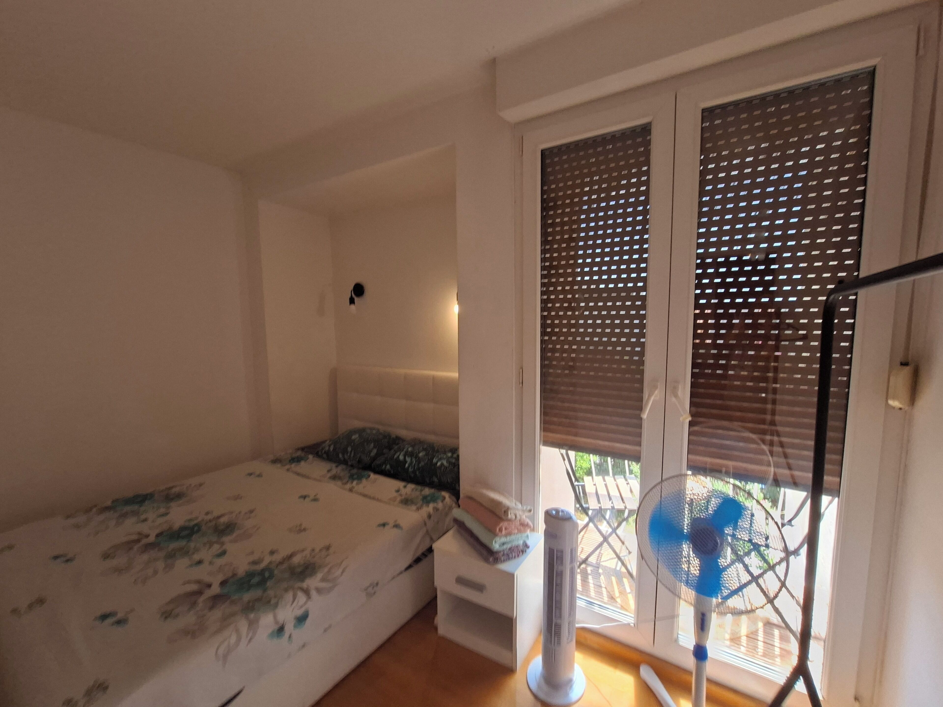 A bedroom with a bed, a standing fan, and a window with blinds.