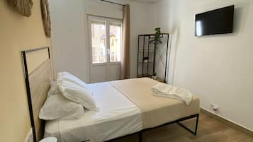 Un dormitorio con cama, ventilador de techo, televisor, mesita de noche con control remoto y ventana con cortinas.