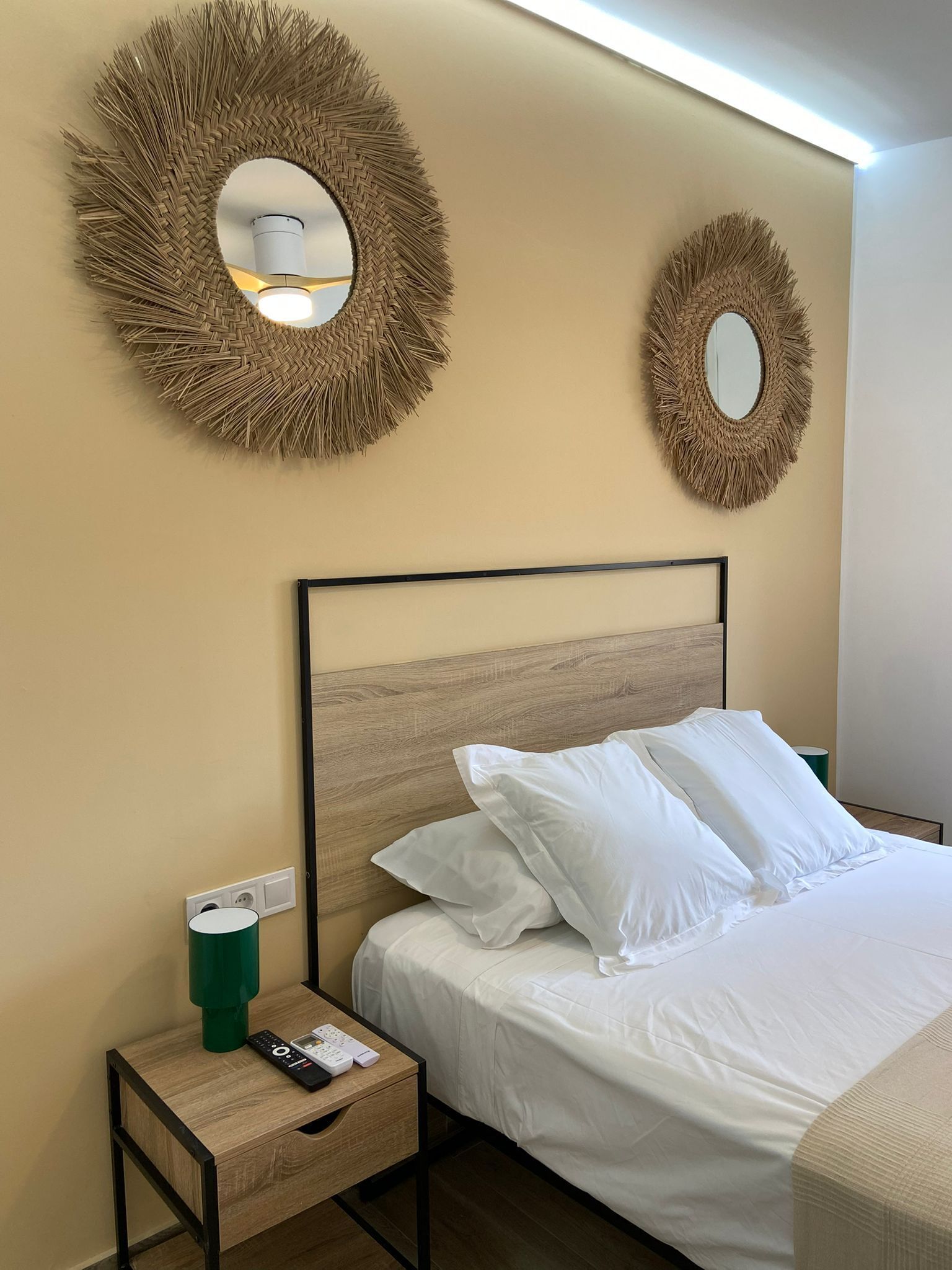Una habitación de hotel con una cama, dos espejos adosados a la pared, una mesita de noche con control remoto y un portavasos verde.