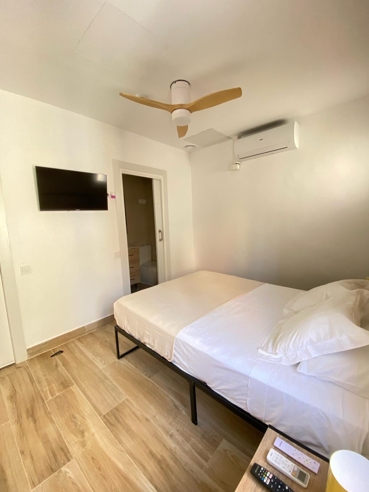 Un dormitorio con una cama, ventilador de techo, equipo de aire acondicionado, televisor y una mesita de noche con control remoto y una taza.