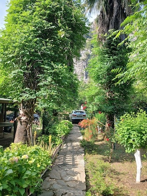 A stone pathway flanked by lush greenery and trees, leading towards a parked car.
