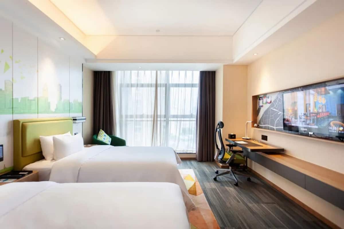 Une chambre d’hôtel avec deux lits, un bureau, une chaise, une télévision et une grande fenêtre avec des rideaux.