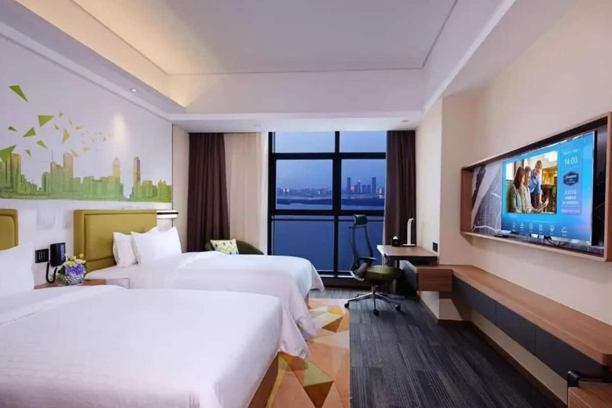 Une chambre d’hôtel avec deux lits, un bureau, une chaise, une télévision et une grande fenêtre donnant sur la ville.