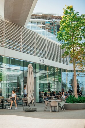 Un bâtiment moderne doté d’une façade en verre, d’un espace extérieur aménagé pour s’asseoir et d’un arbre.