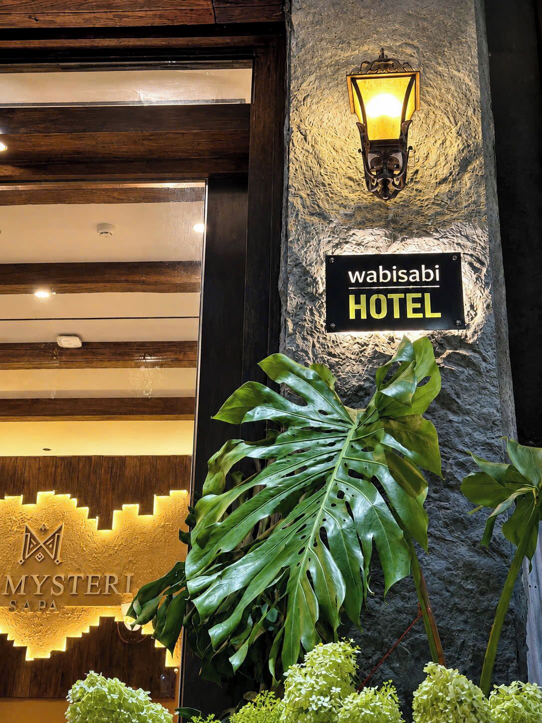 Entrada de un hotel con un letrero que dice 'Wabisabi HOTEL' y una planta grande al frente.