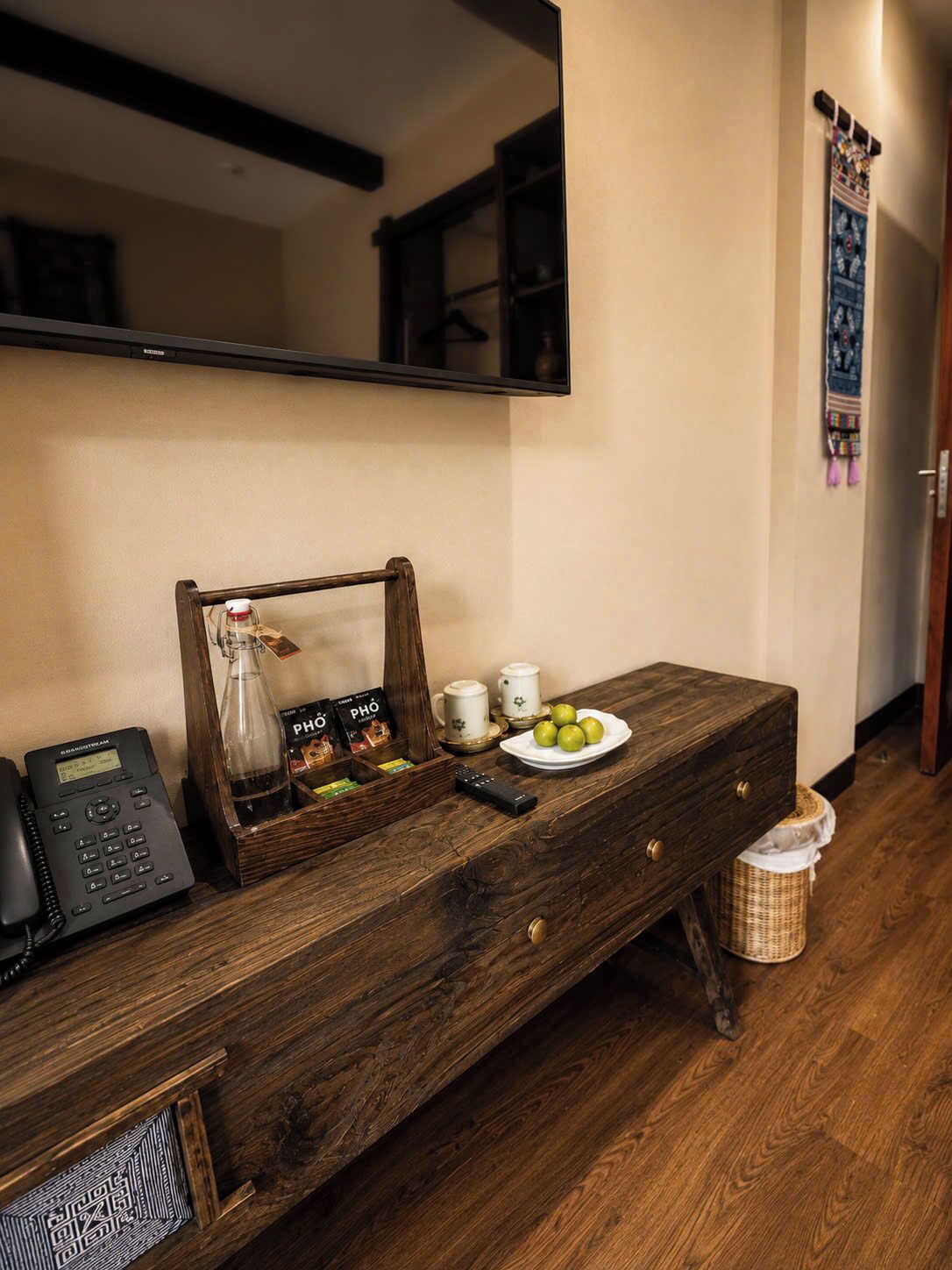 A wooden console table with a telephone, a glass bottle, a tray with coffee cups and a plate of apples, a small wicker basket, and a mirror above.