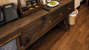 Una consola de madera con un teléfono, una botella de vidrio, una bandeja con tazas de café y una fuente de manzanas, una pequeña cesta de mimbre y un espejo encima.