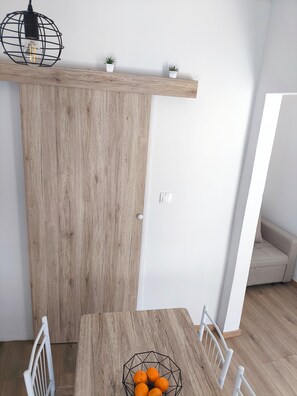 Ruokailutila, jossa on puinen pöytä, tuoleja, geometrisesti muotoiltu kulho täynnä appelsiineja, riippuvalaisin ja valkoinen seinä.