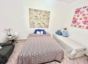 A bedroom with a bed, a small round table with a lamp, a potted plant, and a wall with a floral painting. - Spacious and bright apartment in the center of Torre dell'Orso with private parking space (Torre dell'Orso)