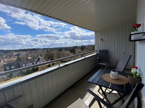 Ein Balkon mit Blick auf eine Stadt, ein Tisch mit Stühlen, ein Blumentopf und ein an der Wand befestigter Fernseher.