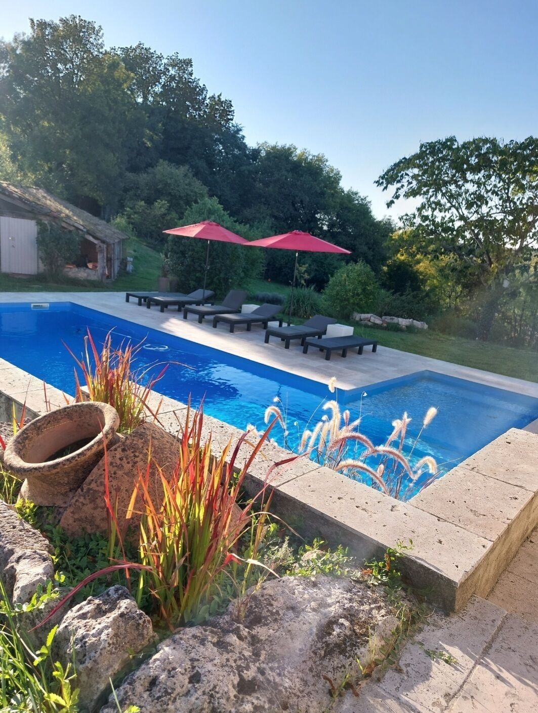 Una piscina con sillas de descanso y sombrillas rojas, rodeada de follaje y un muro de piedra.