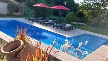 Una piscina con sillas de descanso y sombrillas rojas, rodeada de follaje y un muro de piedra.