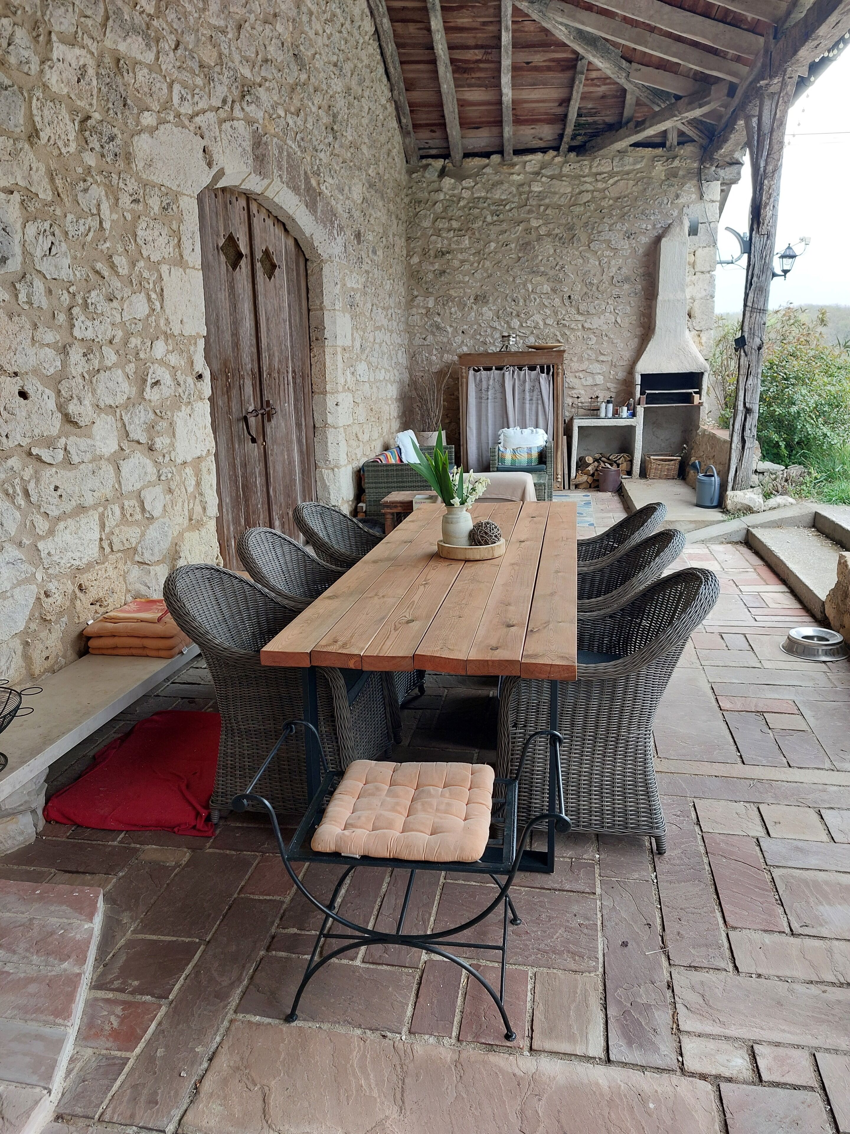Un espacio de comedor al aire libre con paredes de piedra, una mesa de madera y sillas de mimbre, un banco pequeño y un horno de leña.