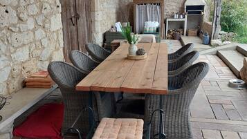 Un espacio de comedor al aire libre con paredes de piedra, una mesa de madera y sillas de mimbre, un banco pequeño y un horno de leña.