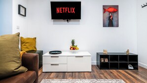 Uma sala de estar com um sofá de couro marrom, uma mesa de centro branca com frutas, uma TV preta na parede e um móvel de estante preto.