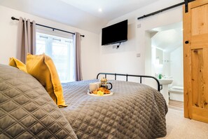 A bedroom with a bed, a bedside table with a cup and saucer, a tray with food, a TV on the wall, and a bathroom visible through an open door. - Unique Devon Cottages - Sunset Cottage (Exmouth)