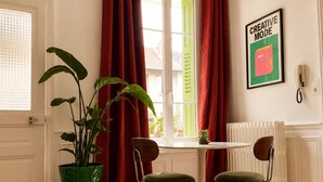 Ein Zimmer mit einem runden Tisch, zwei Stühlen, einer Topfpflanze, roten Vorhängen, einem Heizkörper und einem gerahmten Bild an der Wand.