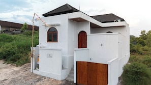 A small, white building with a wooden door and window, surrounded by greenery.