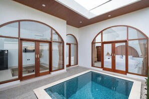 A modern hotel interior with a pool, large arched glass doors, and a visible bedroom with a bed and curtains.