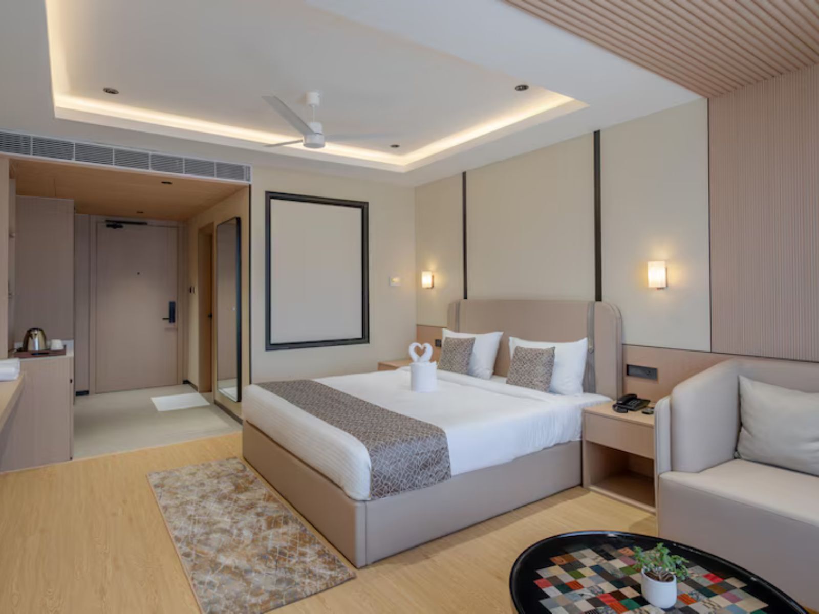 A modern hotel room with a large bed, bedside tables, a sofa, a small table with a plant, and a ceiling fan.