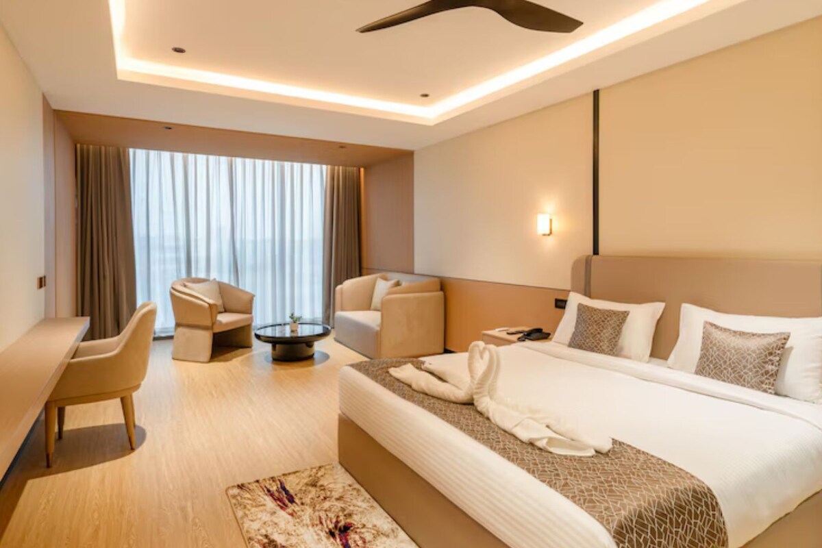 A modern hotel room with a bed, two armchairs, a small table, and a ceiling fan.