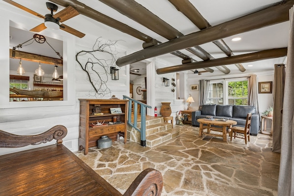 A rustic interior with exposed wooden beams, a stone floor, a wooden staircase, a dining table, a sofa, and a fireplace.