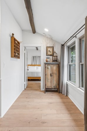 A hallway with wooden flooring, a bed with white bedding, a wooden cabinet with decorative items, and a window with curtains.