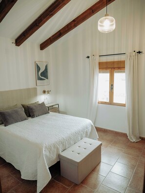 A bedroom with a large bed, a bedside table, a hanging light fixture, a window with curtains, and a wall-mounted artwork.