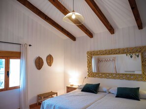 Une chambre Ă coucher avec un lit, deux oreillers, une table de chevet, une lampe, un miroir avec un cadre ornĂ© et une fenĂȘtre avec des rideaux.