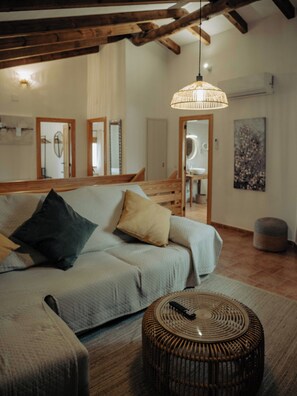 A room with a light-colored sofa, a round wicker ottoman, a hanging light fixture, wooden beams, and a mirror. - Vrbo Property (La Gila,Alcalá del Júcar)