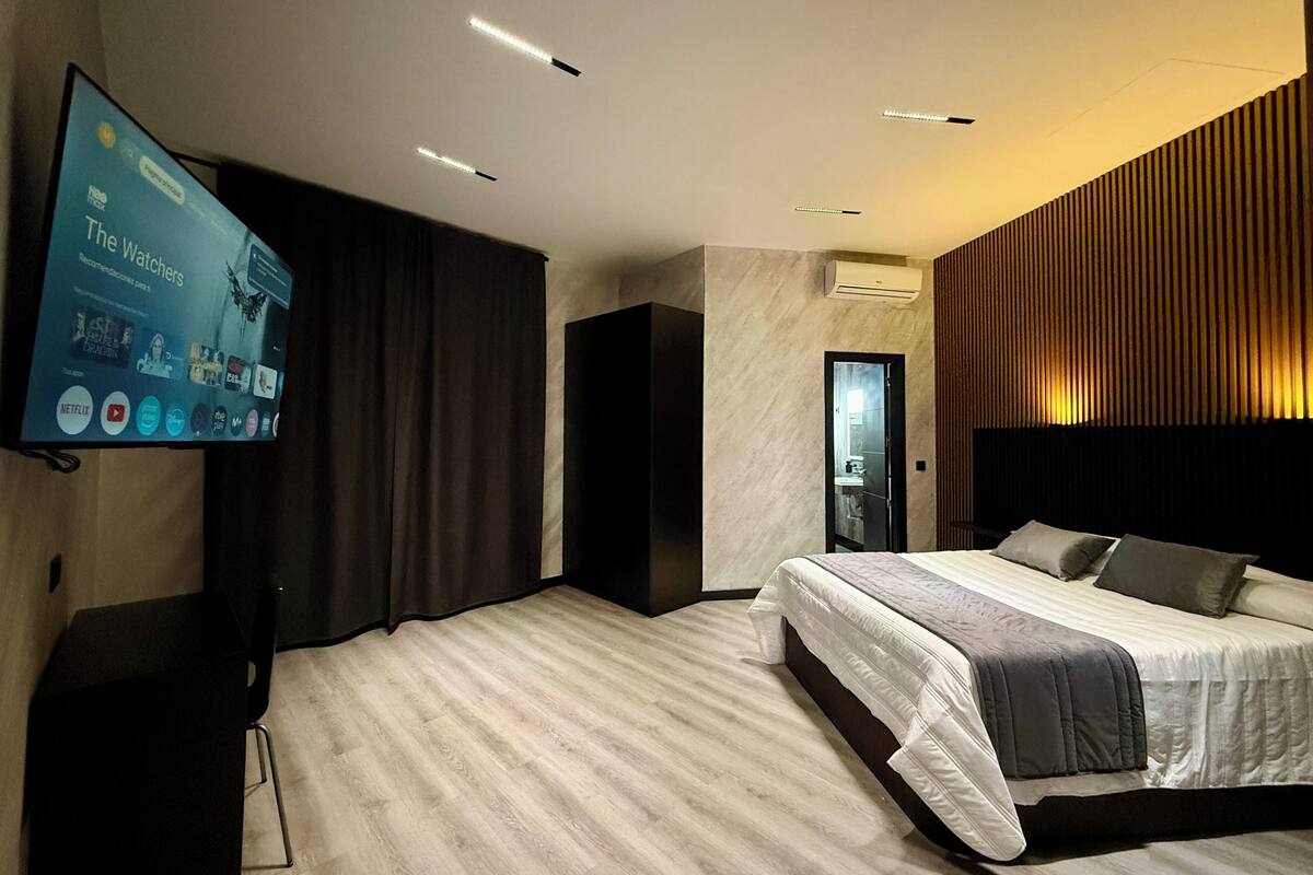 A modern hotel room with a large flat-screen TV, a bed with a headboard, a dark-colored wardrobe, and a wooden panel wall.