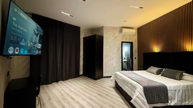 Una habitación de hotel moderna con una televisión de pantalla plana grande, una cama con cabecera, un armario de color oscuro y una pared con paneles de madera.