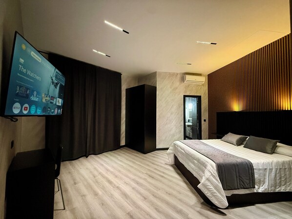 A modern hotel room with a large flat-screen TV, a bed with a headboard, a dark-colored wardrobe, and a wooden panel wall.