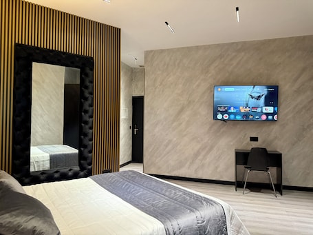 A modern hotel room with a large bed, a TV mounted on the wall, a desk, and a mirror with vertical wooden slats.. Hotel Reset