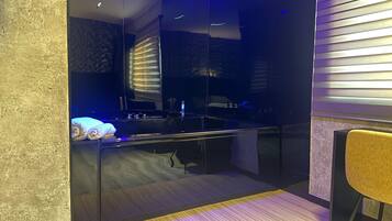 Una habitación moderna con una cama, una mesita pequeña y una silla. Hay una ventana con persianas, y el suelo es de madera.