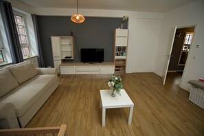 Un salon avec un canapé, un téléviseur, une table blanche ornée d’un vase, un plancher en bois et un mur gris.