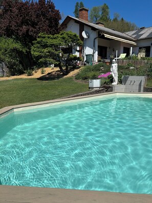 Una piscina cristalina con una estatua, rodeada de un césped cuidado y una casa con chimenea de ladrillo.