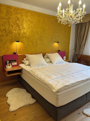 A bedroom with a large bed, two bedside tables, a chandelier, and a fluffy rug. - Golden Rooms Exclusive (Völkermarkt)